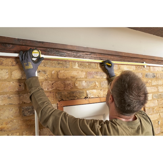 STANLEY 30-710 tape measure used by a person in gloves to measure above a door frame on a brick wall.