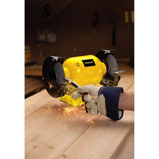 STANLEY STGB3715 bench grinder in use on wooden workbench, sparks flying as gloved hand grinds metal tool.