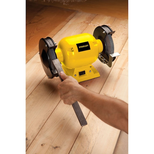 Photo of STANLEY STGB3715 bench grinder in use on wooden surface, yellow body, metal grinding wheel.
