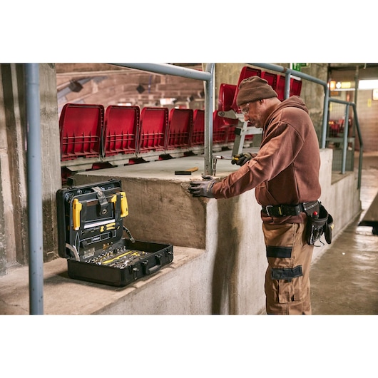 STANLEY STMT98109-LA tool set in open case on concrete ledge, worker using tools, red stadium seats in background.
